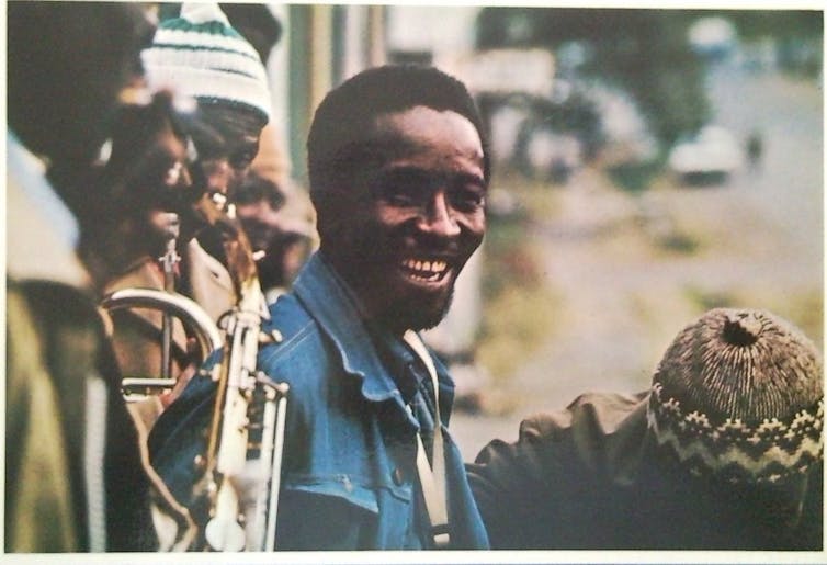 A bearded man is laughing as he stands in a row with musicians on brass instruments.