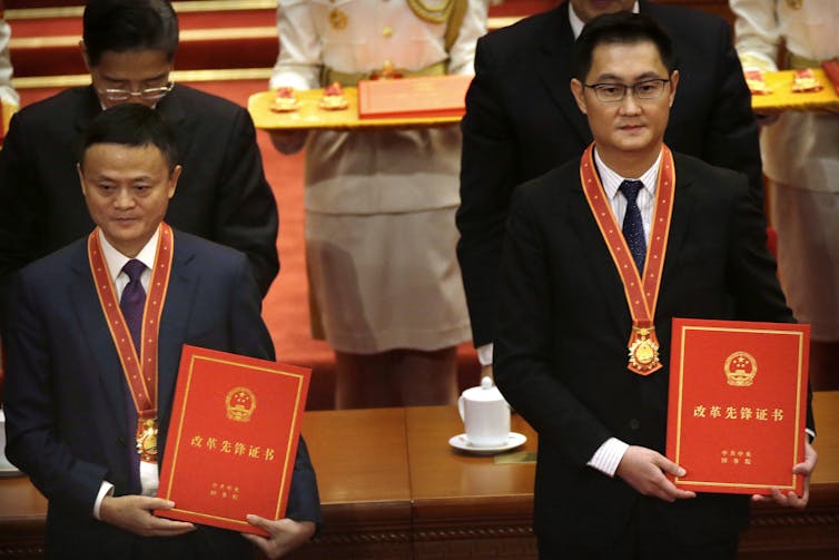 Two men in suits stand with red sashes around their necks and hold a red book.