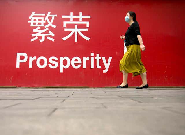 A woman in a yellow skirt and black shirt wearing a mask walks past a large red banner reading 'prosperity' in English and Chinese