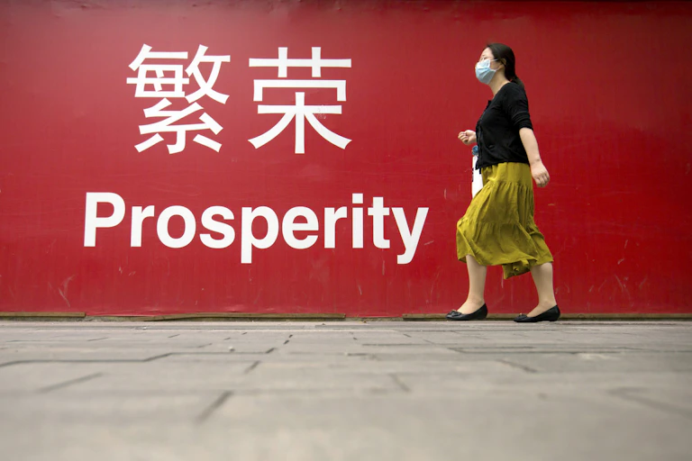 A woman in a yellow skirt and black shirt wearing a mask walks past a large red banner reading 'prosperity' in English and Chinese