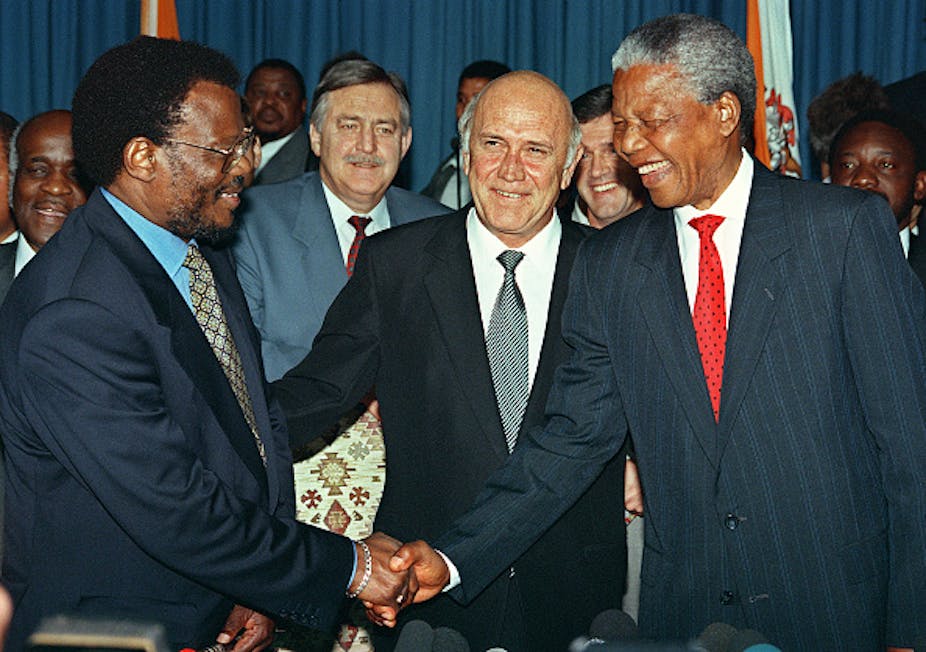 A man smiles as he is flanked by two others shaking hands, also smiling. All of them wearing suits.