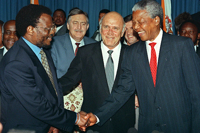 A man smiles as he is flanked by two others shaking hands, also smiling. All of them wearing suits.