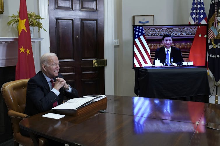 A smiling man sits at his desk with his hands clasped while another man appears on a video screen to his left.
