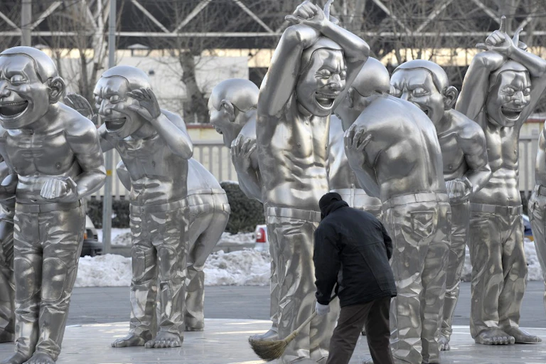 Statues argentées monochromes d'hommes torses nu grimaçant balayées par un homme de dos.