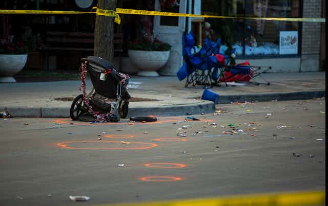 A mangled stroller and camp seats line the street with yellow police tape fluttering above.
