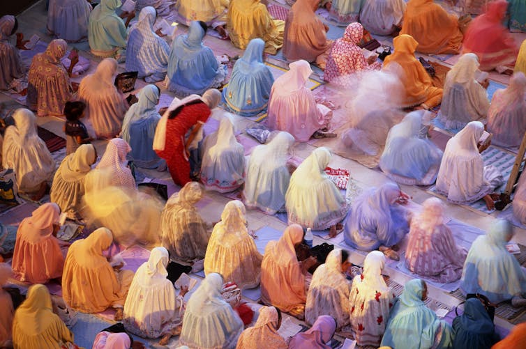 Rows of Muslim women, wearing headscarfs, seated in a prayer position.