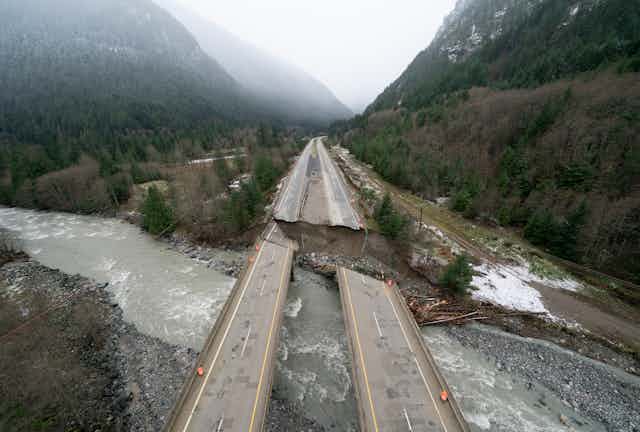 A portion of a highway between two tall mountains is destroyed. A raging river runs under the portion that is still intact.