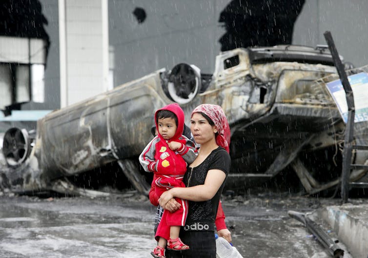 A woman stands holding her child in front of a blown-up, flipped-over car