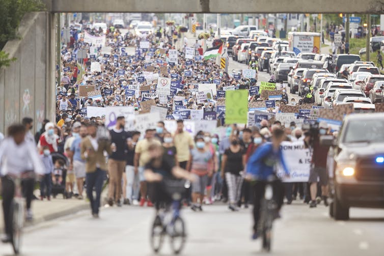 A large crowd of people is seen marching on the street, next to a line of cars.