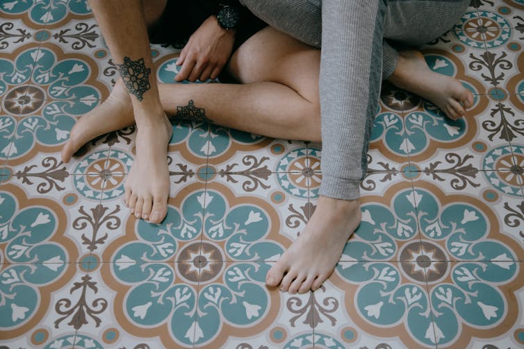 Two young people without shoes sit on a tiled floow.