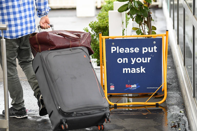 A sign that reads "Please put on your mask" is pictured alongside a person in a checkered blue and white shirt and grey pants pulling two suitcases.