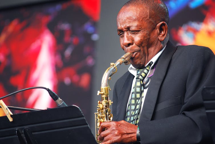 An elder man in a suit and tie blows into a saxophone on stage, bright lights behind him.