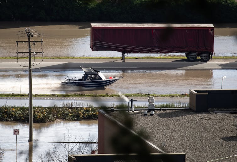 A motorboat travels along a highway flooded with brown water past an abandoned transport truck.