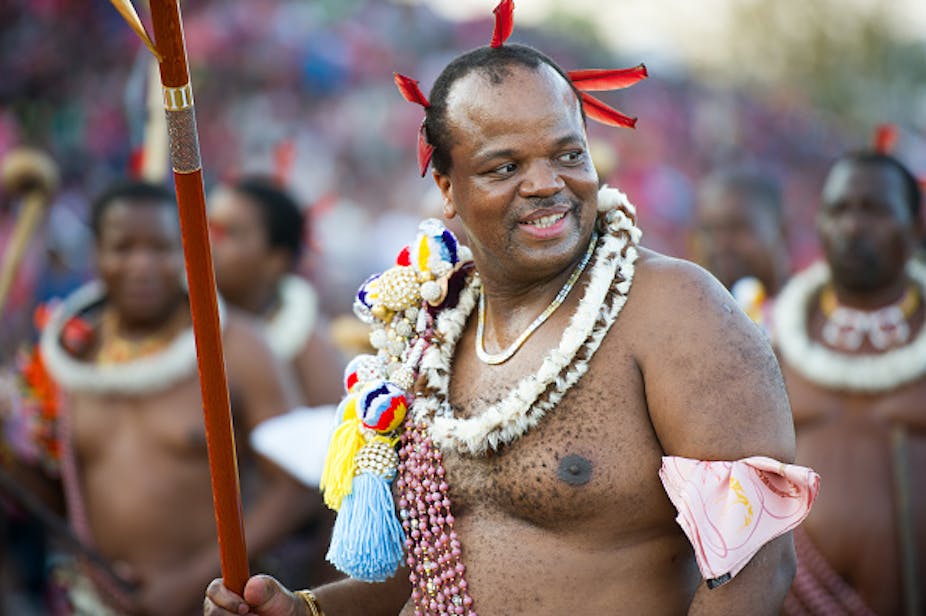 King Mswati II dressed in traditional Swazi king, carrying a sceptre, with his hair decorated with feathers, smiles at the camera