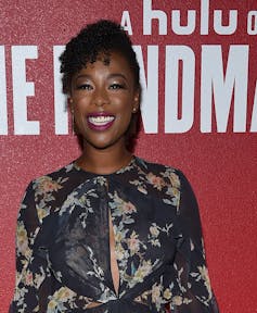 A Black woman dressed glamorously in red lipstick is seen arriving at an event in front of a Hulu / Handmaid's Tale sign.