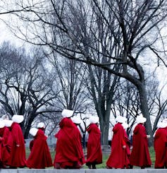 A group of women in red cloaks and bonnets are seen walking by a cluster of trees outside.