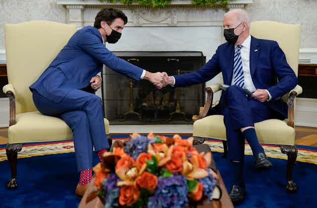 A man with dark hair in a blue suit wearing a mask shakes the hand of a man with grey hair in a blue suit wearing a mask in an ornate office with autumn flowers in front of them.