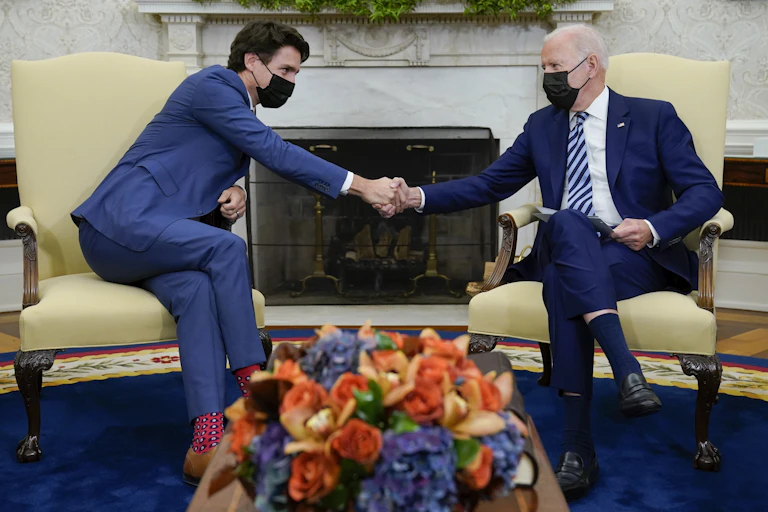A man with dark hair in a blue suit wearing a mask shakes the hand of a man with grey hair in a blue suit wearing a mask in an ornate office with autumn flowers in front of them.