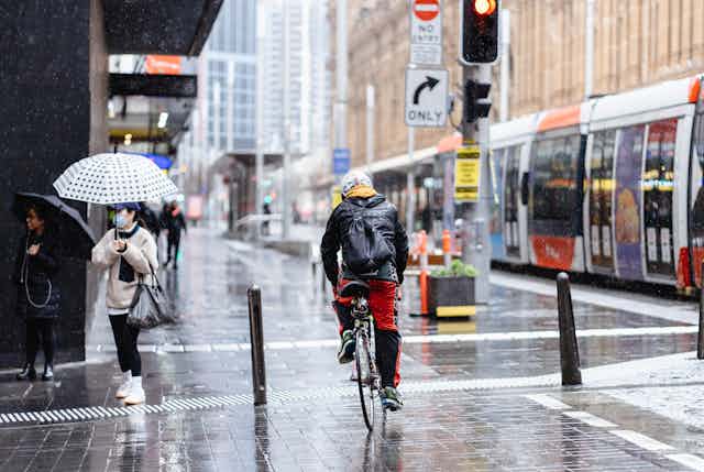 Cyclist going through city on rainy day, with a tram on his right and a pedestrian on the left holding an umbrella