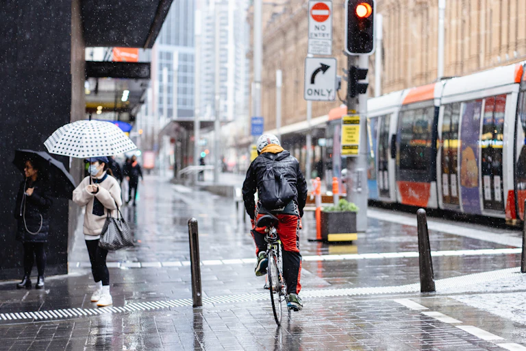 Cyclist going through city on rainy day, with a tram on his right and a pedestrian on the left holding an umbrella