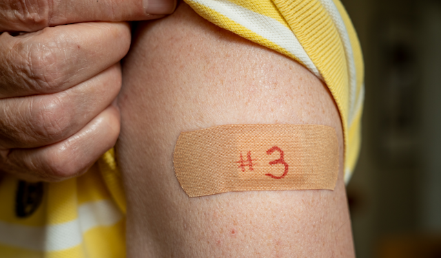 An older man holds up his shirt sleeve to reveal a bandaid on his arm with the number three written on it.