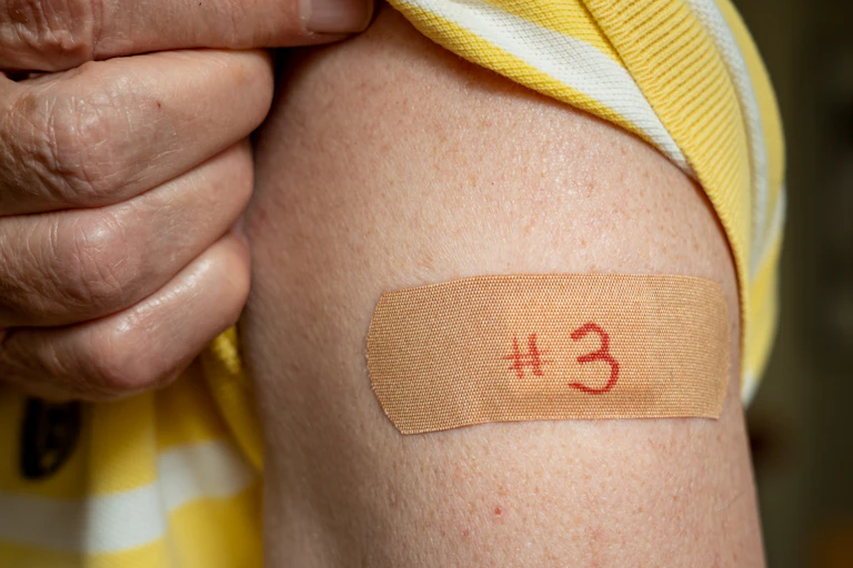 An older man holds up his shirt sleeve to reveal a bandaid on his arm with the number three written on it.