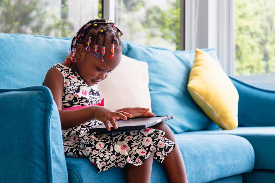A young girl sitting on the sofa and playing on tablet in the living room.