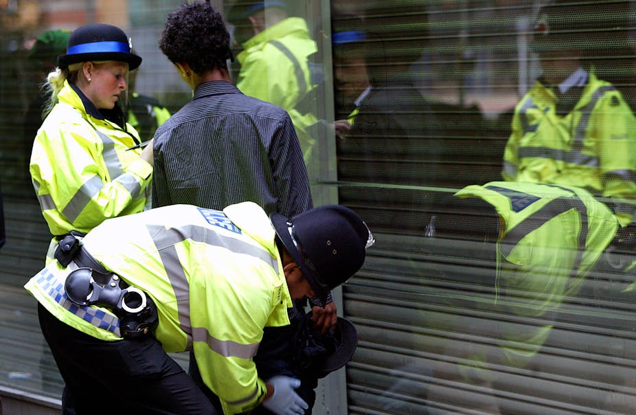 Two police officers in flourescent jackets searching a young black man in Manchester. all are facing away from the camera.