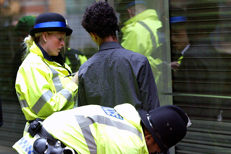 Two police officers in flourescent jackets searching a young black man in Manchester. all are facing away from the camera.