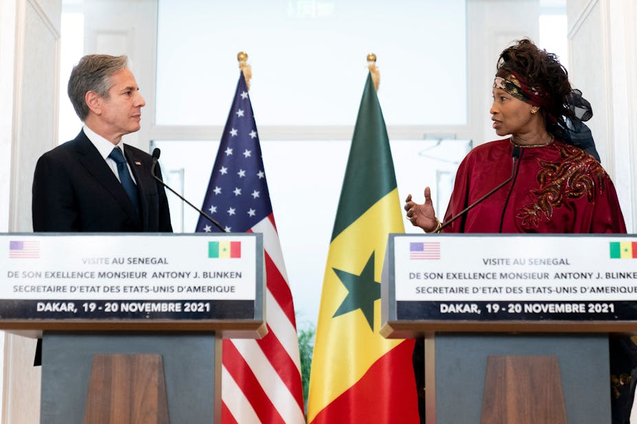 A man and a woman speak while looking directly at each other. They have national flags behind them.