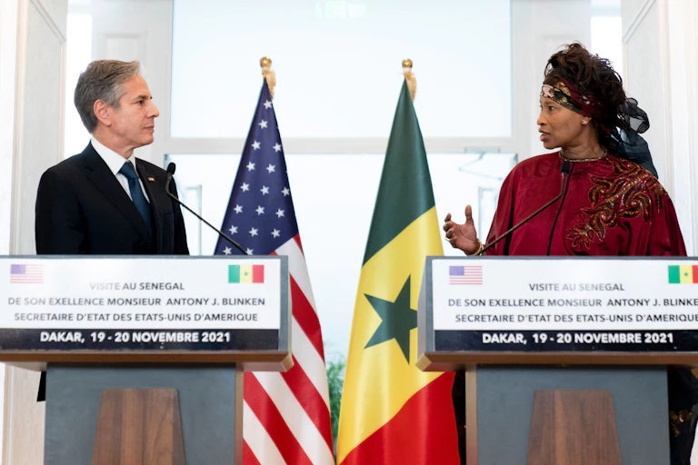 A man and a woman speak while looking directly at each other. They have national flags behind them.