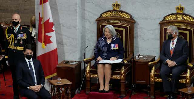 A woman with a grey bob, blue dress with ceremonial badges reads from a speech while sitting on a throne that has ER etched into it. A man with a mask with grey hair sits to her left, a man in a blue suit with clasped hands wearing a mask sits to her right. 