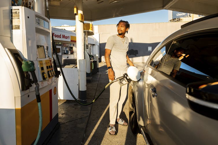 A man in a grey T-shirt and pants pumps gas into his car.