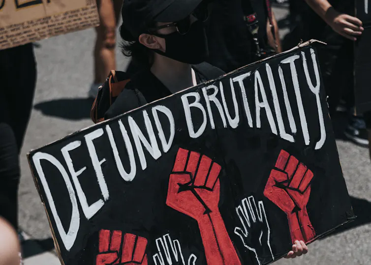 A person wearing a face mask carries a sign that reads 'defund brutality'