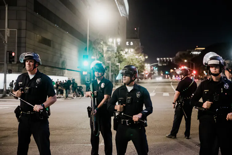 A group of police officers stand together wearing helmets with faceshields above their heads.