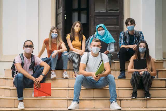 A group of college students wearing masks sit on the steps of a building.