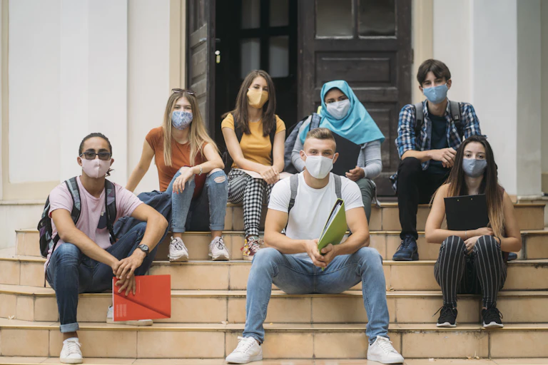 A group of college students wearing masks sit on the steps of a building.