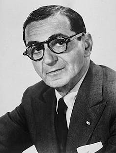 Black and white photo of a man in a suit, tie and glasses.
