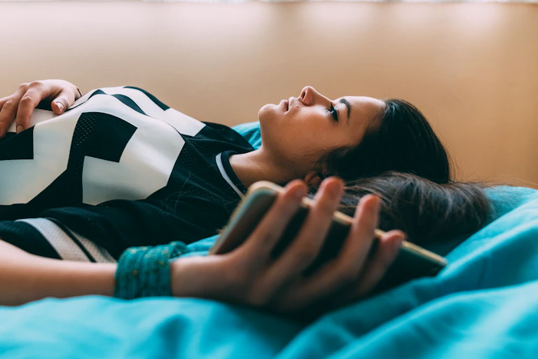 A sad young woman lies on a bed with phone in hand.