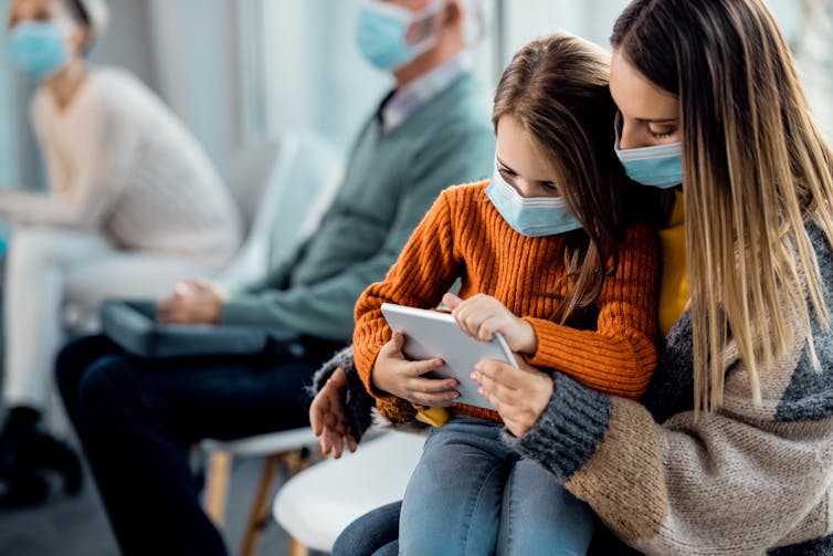 Worried girl sits on her mother's lap, looking at a tablet.