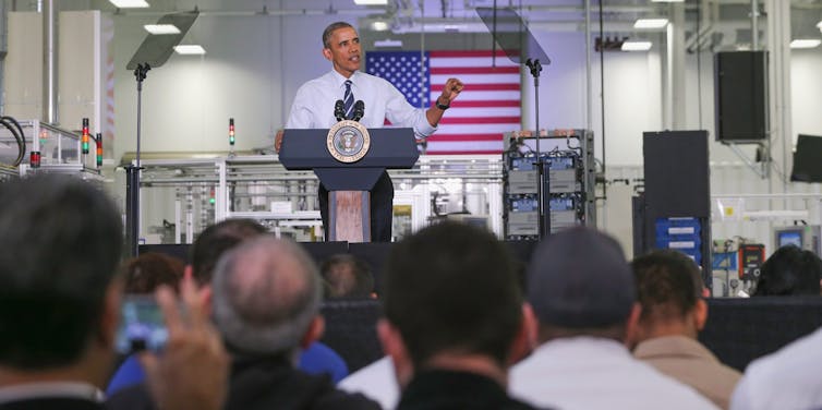 A man stands at a podium with an American flag behind him talking to factory workers.