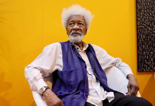 An elderly man sits in front of a bright yellow wall. He wears a purple waistcoat and has a white beard and Afro. He looks into the distance, slightly pensive.