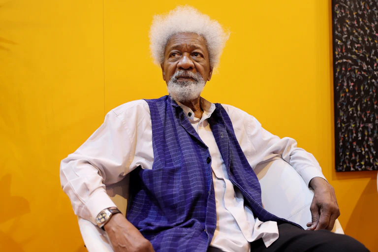 An elderly man sits in front of a bright yellow wall. He wears a purple waistcoat and has a white beard and Afro. He looks into the distance, slightly pensive.