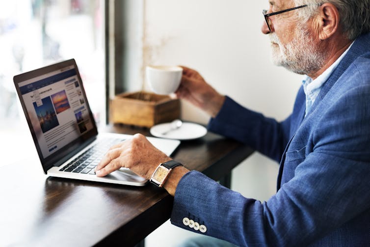 an older man drinks from a teacup while sitting in front of a laptop