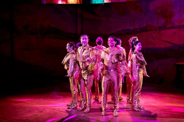Production image: a cast dressed in gold, lit in pink