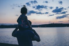 Ein kleines Mädchen sitzt auf den Schultern ihres Vaters, während sie einen Sonnenuntergang beobachten.