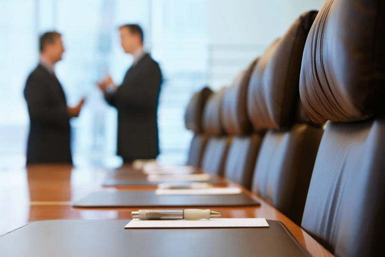 Two white men stand at the end of a boardroom table with leather chairs in the foreground.