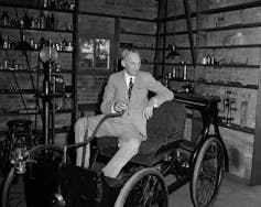 A man in a pale suit and dark tie sits on an antique car.