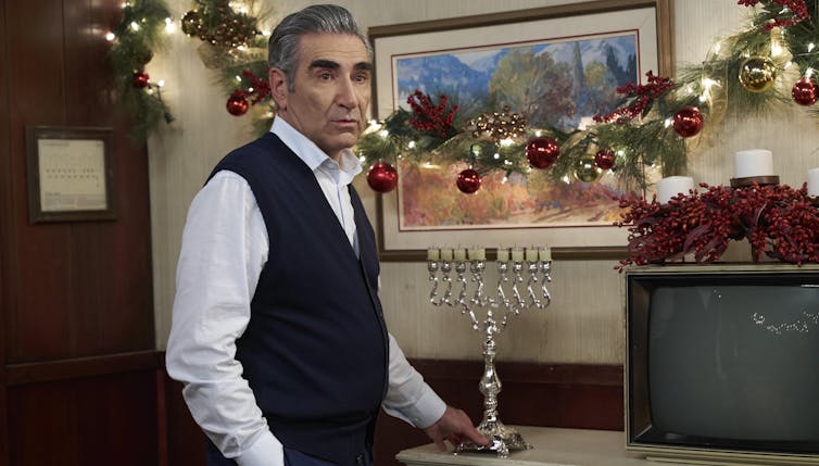 A man in a shirt and vest is seen with a menorah in front of Christmas evergreen boughs and lights on the wall.
