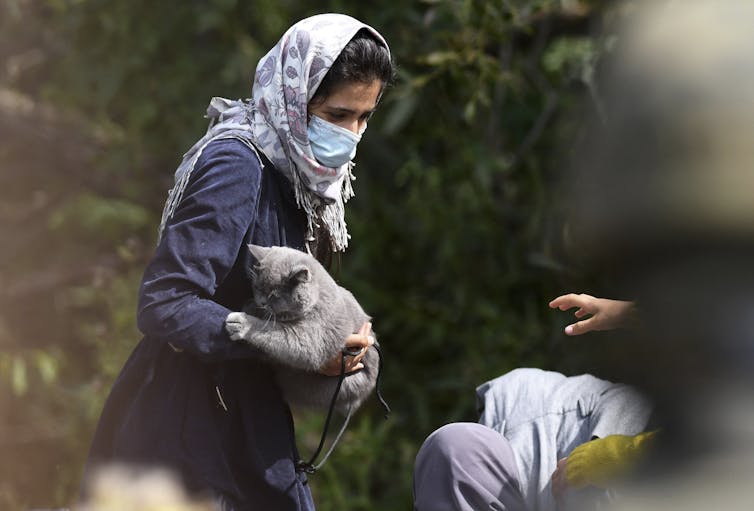 A woman in a head kerchief and wearing a mask holds a grey cat.